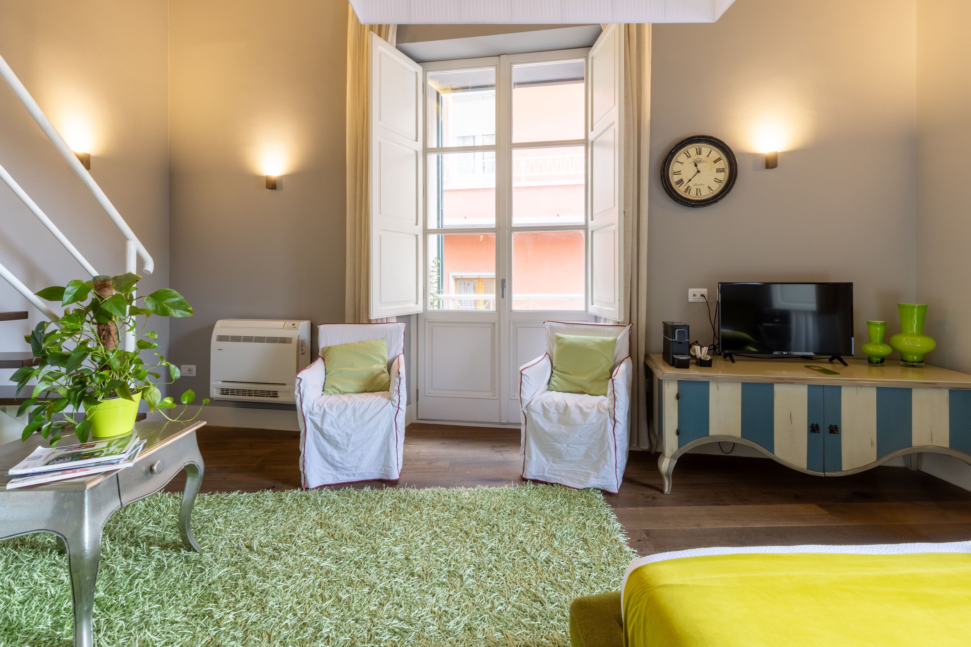A room with a bed, a desk with a TV, two chairs, a window, and a wall clock.