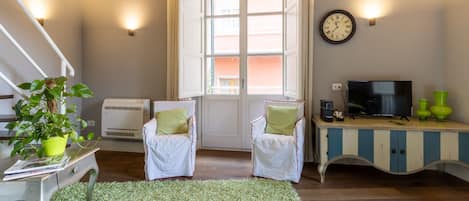 A room with a bed, a desk with a TV, two chairs, a window, and a wall clock.