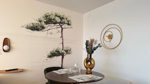 Un coin repas comprenant une table ronde, deux chaises, un vase avec des fleurs séchées et une horloge murale.