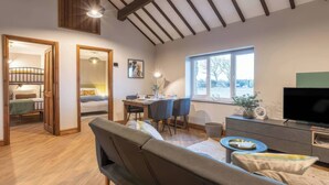 A cozy living room with a sofa, a dining table, and a TV. There's a window with a view of the outside. - Blue Bird Cottage (Gaerwen)
