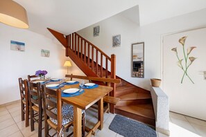 A dining area with a wooden table set for four, a staircase, and a door with a floral painting. - Holiday Home "Ty Gwen - Plouezoc'h" with Sea View and Wi-Fi (Plouezoc'h)