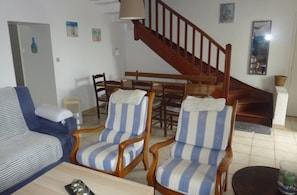Een woonkamer met een bank, twee gestreepte fauteuils, een eettafel met stoelen, een houten trap en een spiegel aan de muur.