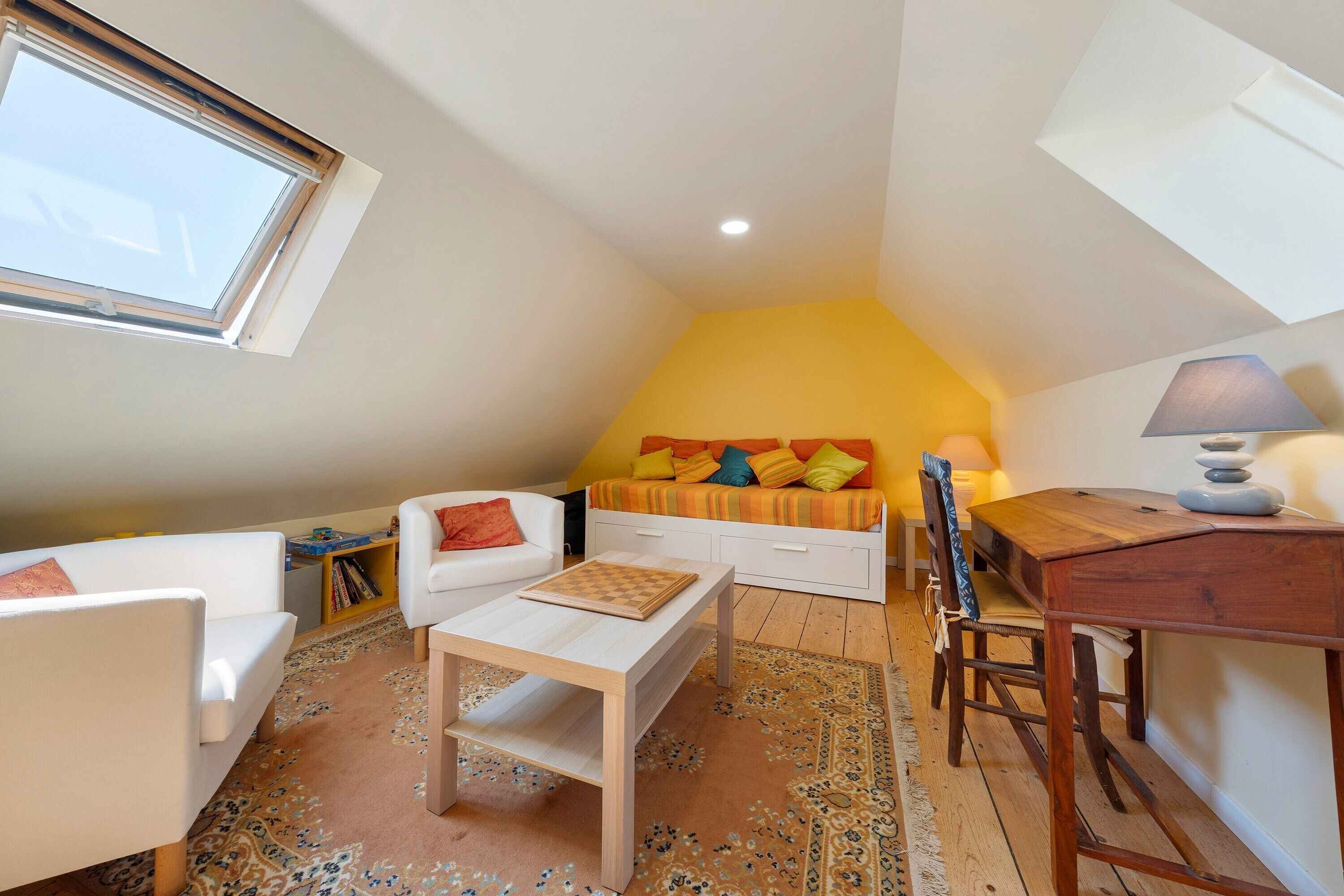 A cozy loft bedroom with a sloped ceiling, a bed with colorful bedding, a white sofa, a small wooden table, and a lamp.