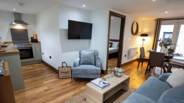 A modern living room with a grey sofa, a wooden coffee table, and a grey armchair. A flat-screen TV is mounted on the wall, and a dining area with a table and chairs is visible. - Beech Cottages (Llanrwst)