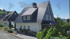 A traditional house with a steeply pitched roof, a well-maintained garden with various plants and flowers, and a clear sky. - Holiday Apartment “Olsberg” with Mountain View, Private Terrace and Wi-Fi (Olsberg)