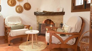 Een knus ogende kamer met een open haard, rieten stoelen, een klein rond tafeltje en een kapstok aan de muur.