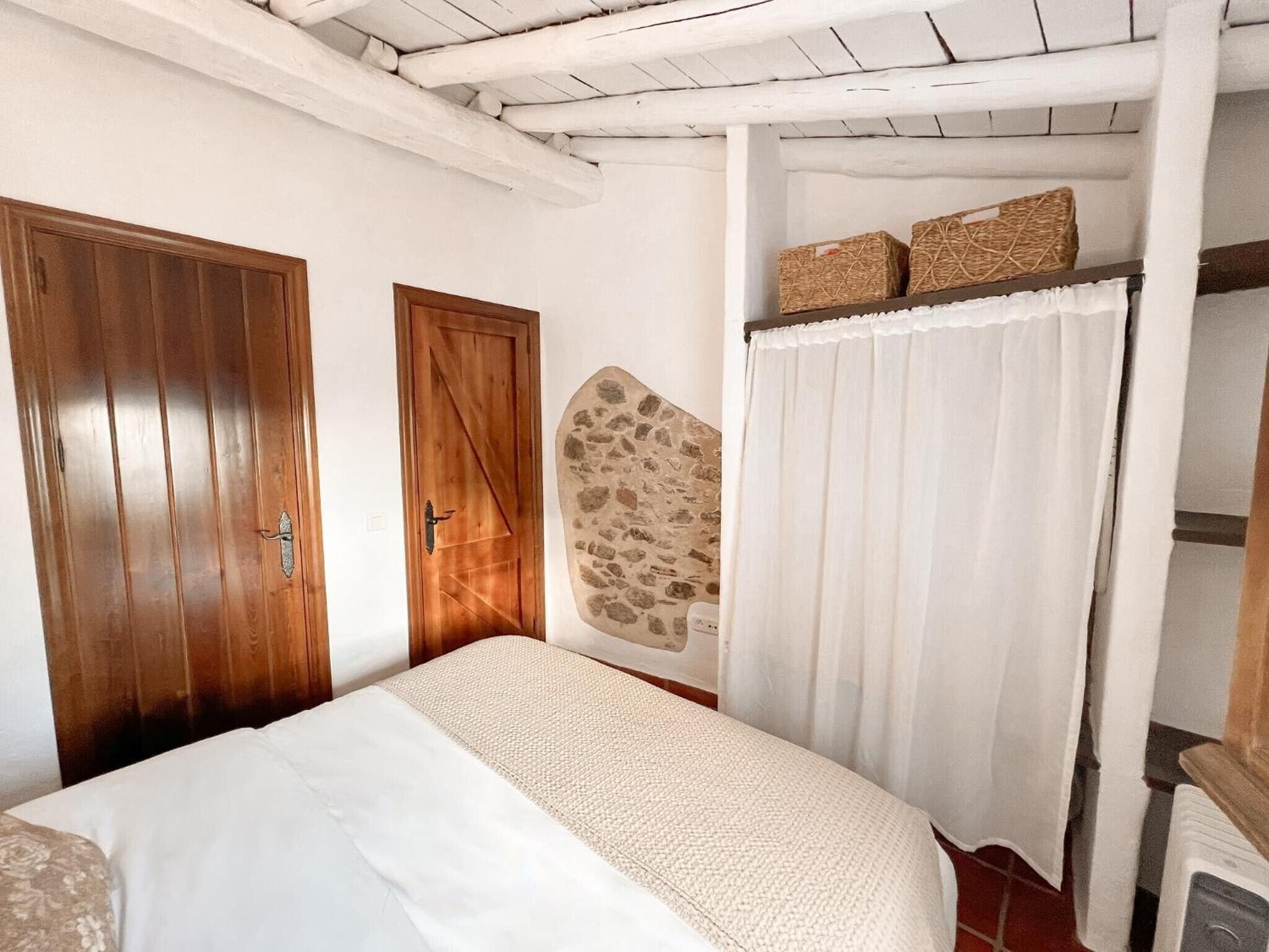 Une chambre à coucher avec un lit, deux portes en bois, un mur en pierre, un rideau blanc et une étagère avec des paniers.