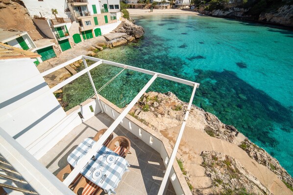 A terrace with a table and chairs, overlooking a clear blue sea and rocky coastline.