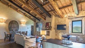 Un intérieur rustique avec des poutres apparentes, un mur en pierre, une cheminée, un coin repas avec une table et des chaises, et une grande fenêtre offrant une vue imprenable.