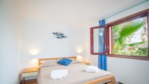 A bedroom with a bed, bedside tables, a nightstand, a window with a view of greenery, and a wall-mounted light fixture. - Waves Apartment, only 250m from the sea  (Tortolì)
