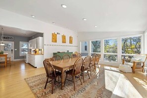 Un comedor amplio con una mesa y sillas de madera, una cocina al fondo y ventanas grandes con vistas a árboles.