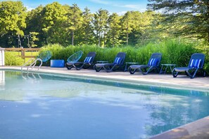 Zona junto a la piscina con sillas de descanso y una pequeña mesa, rodeada de follaje y árboles.