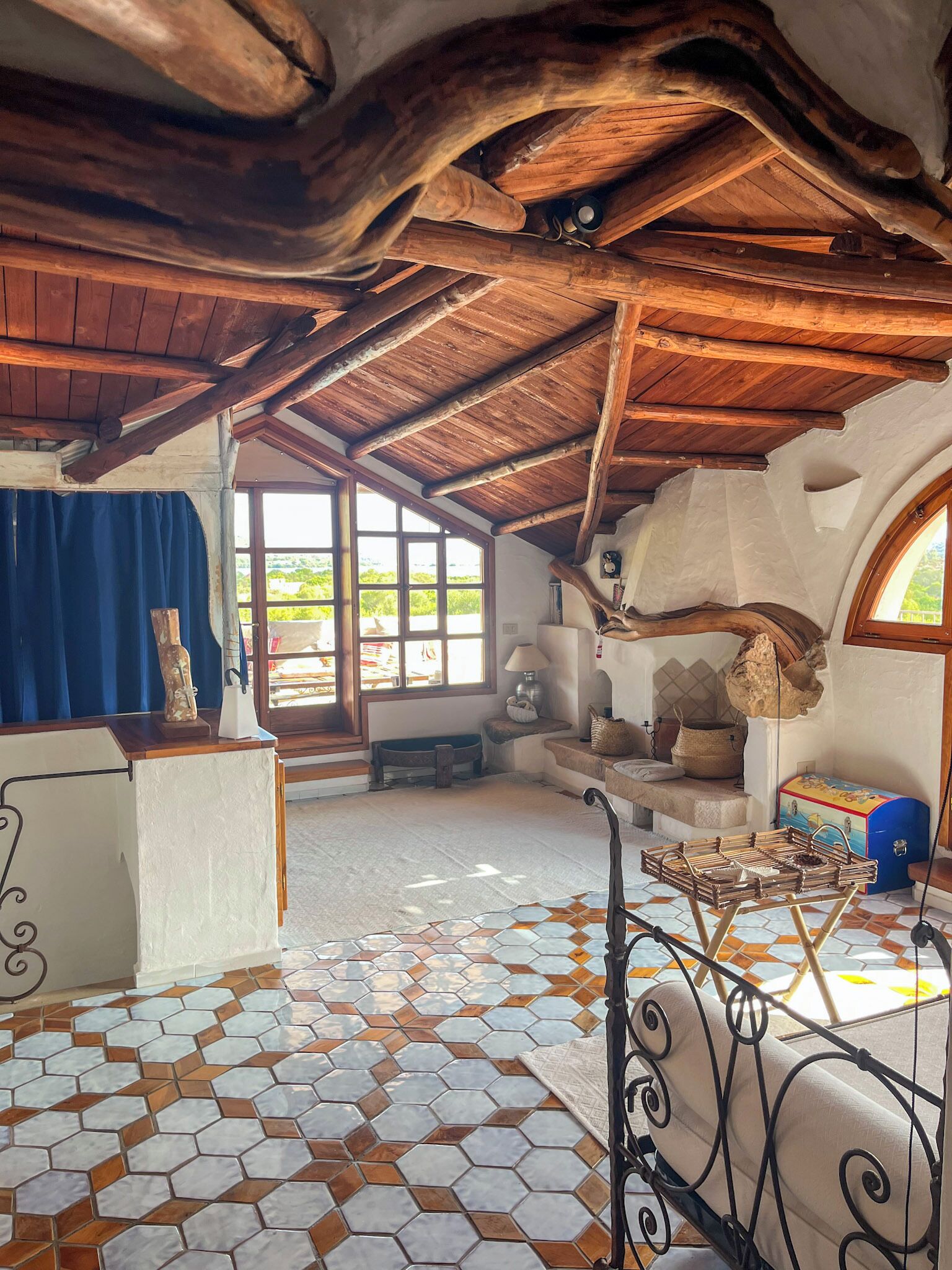 A rustic room with a wooden ceiling, a tiled floor, and a large window offering a view of greenery.
