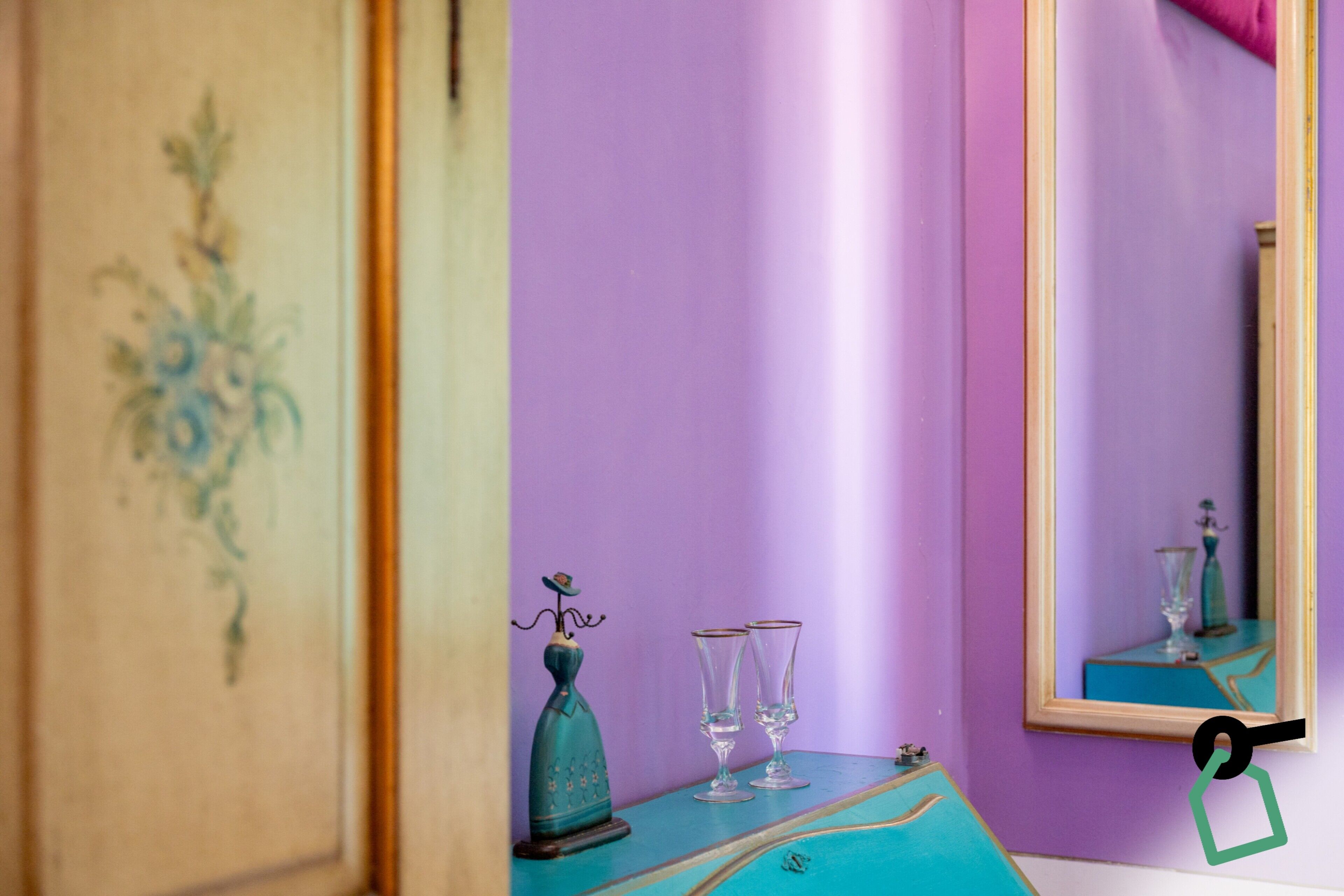 A room with a purple wall, a wooden cabinet with a floral design, a teal table with two glasses and a decorative bottle, and a mirror reflecting a vase and a table.
