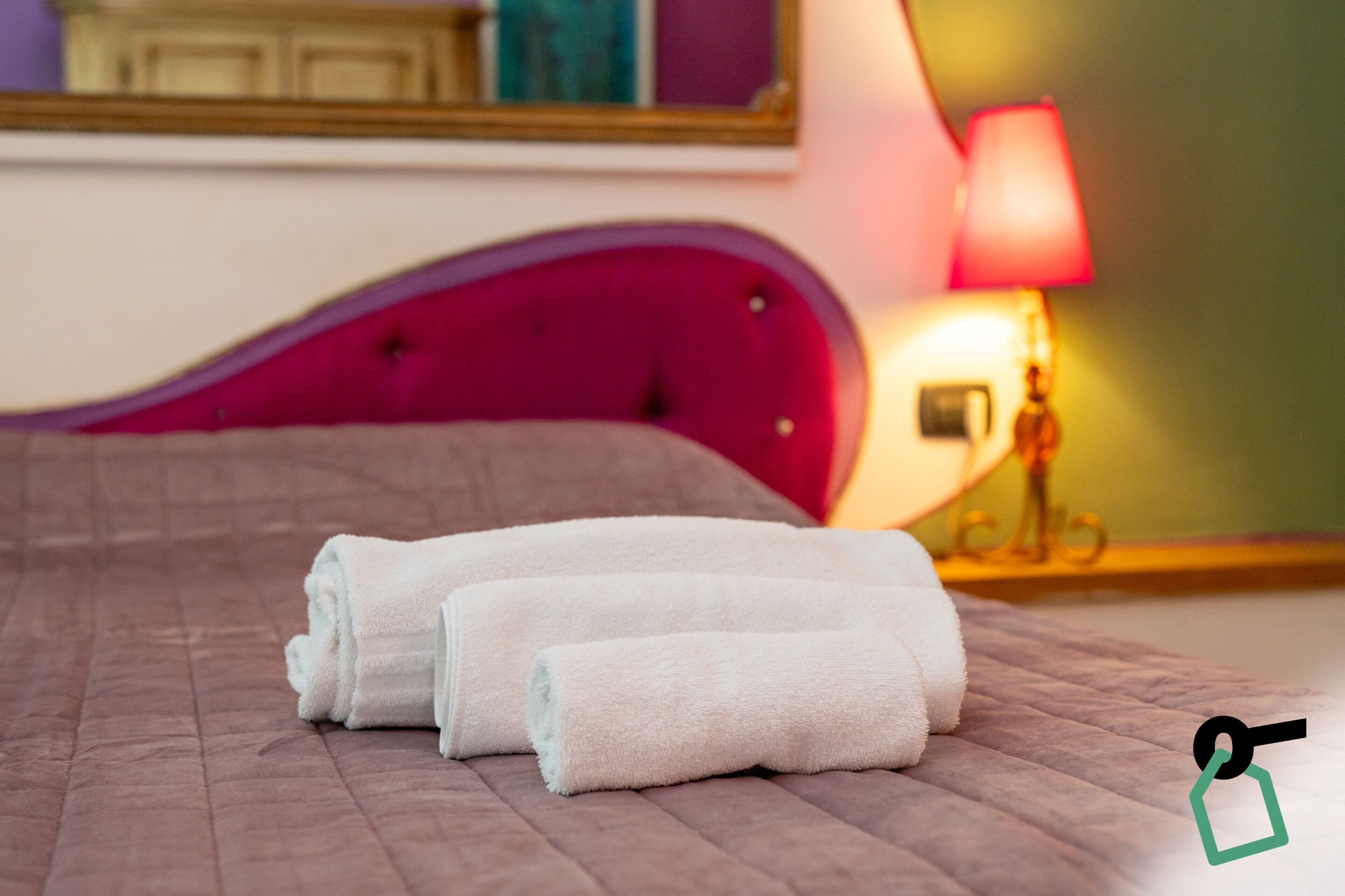 A bed with a pink headboard, two rolled white towels, and a bedside lamp.