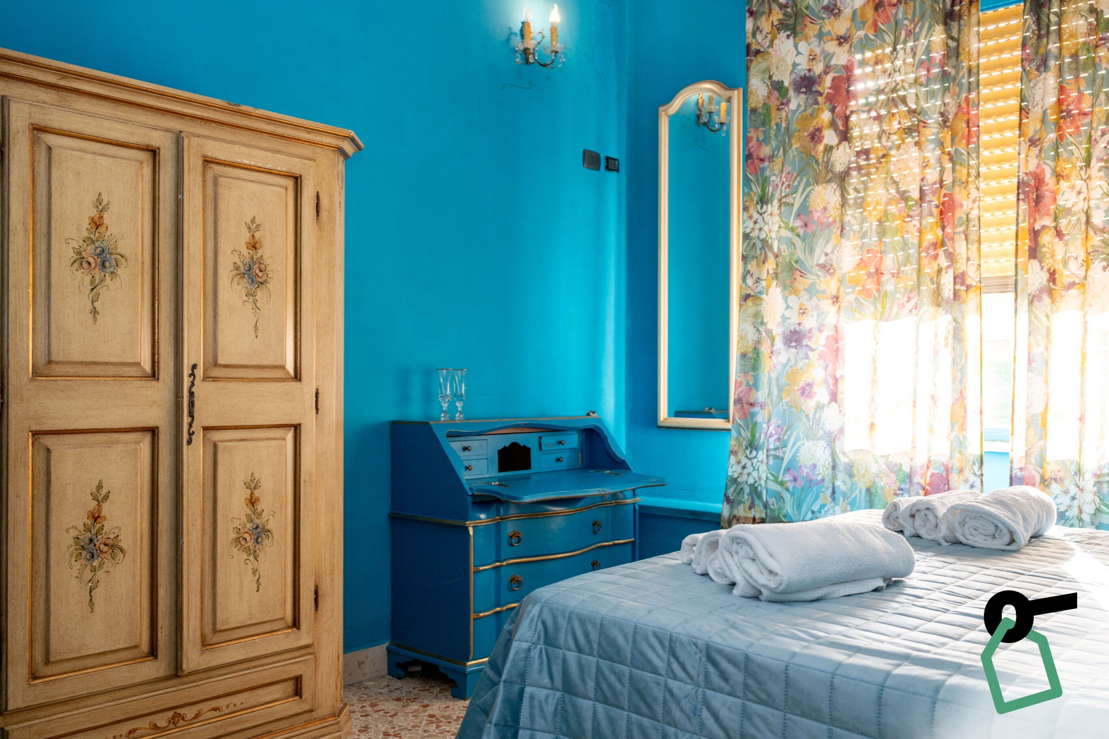 Ein Zimmer mit einem Bett, einem Schminktisch, einem Spiegel und einem Fenster mit floralem Vorhang.