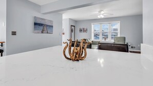 Un salón amplio con una mesa de centro de mármol blanco, un cuadro grande en la pared, un ventilador de techo y vistas a través de una ventana.