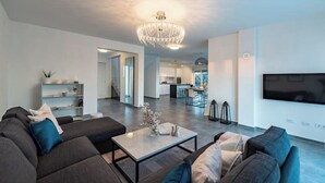 Ein modernes Wohnzimmer mit einer großen Eckcouch, einem Couchtisch und einer Chandelier. Im Hintergrund ist eine Küche mit einem großen, an der Wand befestigten Fernseher zu sehen.