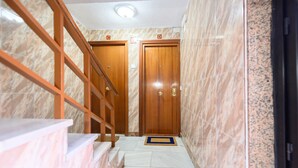 Un couloir au sol de marbre, avec des portes en bois portant les lettres C, D et E, et un escalier en bois sur la gauche.