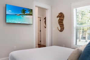 Een slaapkamer met een bed, een tv aan de muur die een strandscène toont, een decoratieve houten zeepaard en een raam met luxaflex.