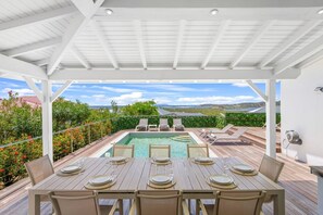 Un espacio al aire libre techado con piscina, mesa de comedor para cuatro personas y sillones de descanso.