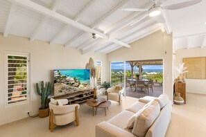 A spacious living room with a large flat-screen TV, comfortable seating, a cactus plant, and a view of the ocean through an open door. - Villa Alanca • Vue Mer & Accès Plage à Pied (Orient Bay)