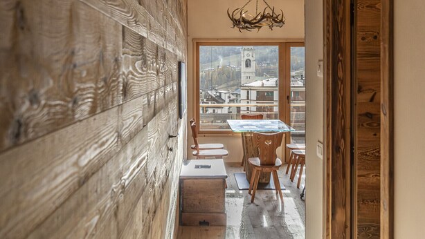 A narrow wooden hallway with a dining area at the end, featuring a table and chairs, and a view of a tower through large windows. - Poligrafica 1 House, quiet and comfortable (Cortina d´Ampezzo)