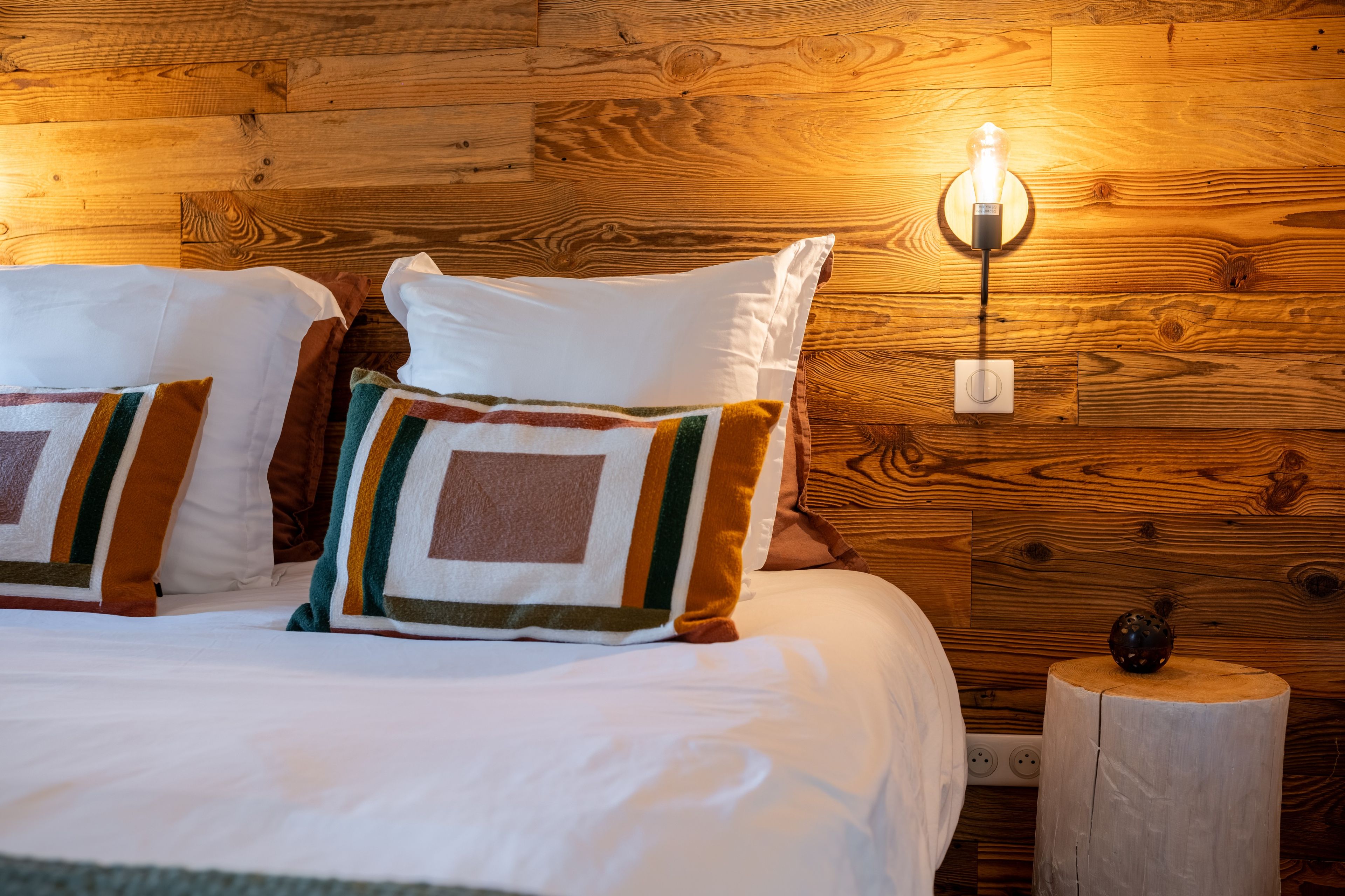 A bed with white bedding and patterned pillows, a wall-mounted light, and a wooden nightstand with a decorative object.