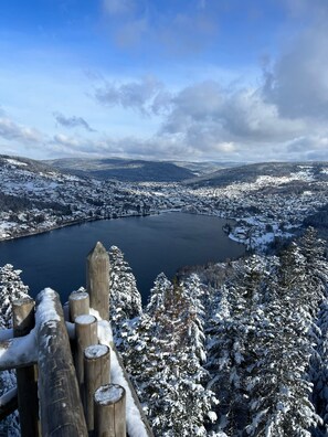 Ein weiter Blick über eine verschneite Landschaft mit einem See, bewaldeten Hügeln und einem Holzzaun im Vordergrund.