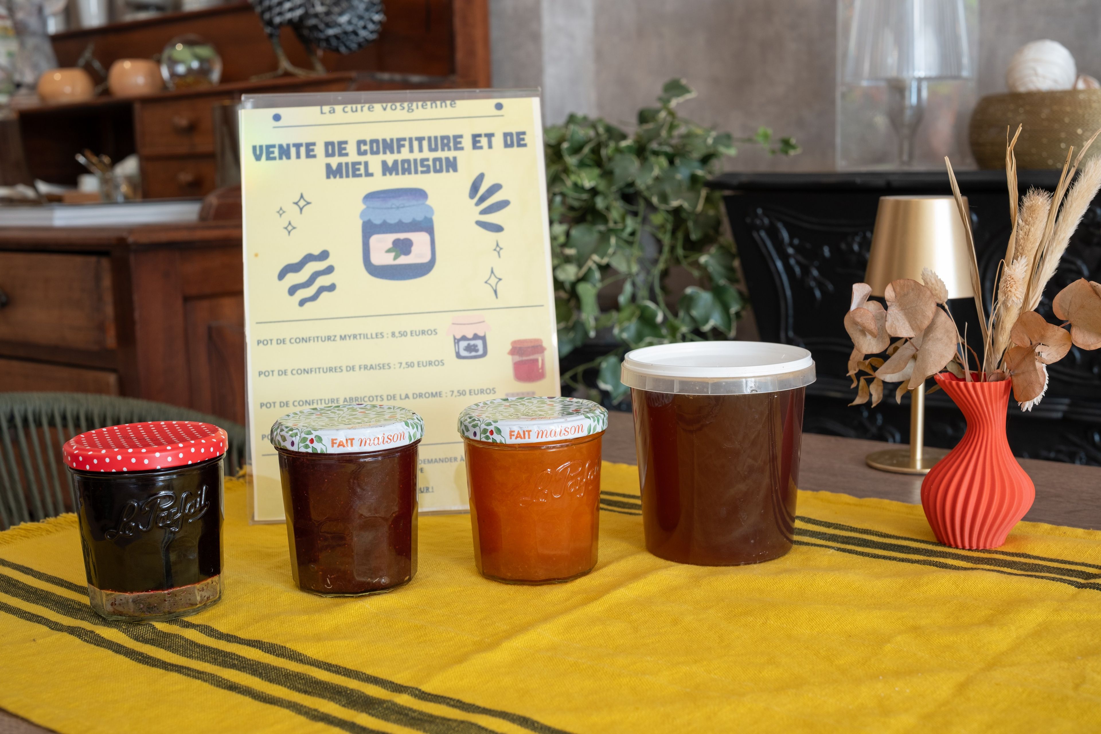Un tavolo con barattoli di conserve, un cartello che pubblicizza marmellate fatte in casa e un vaso con piante essiccate.