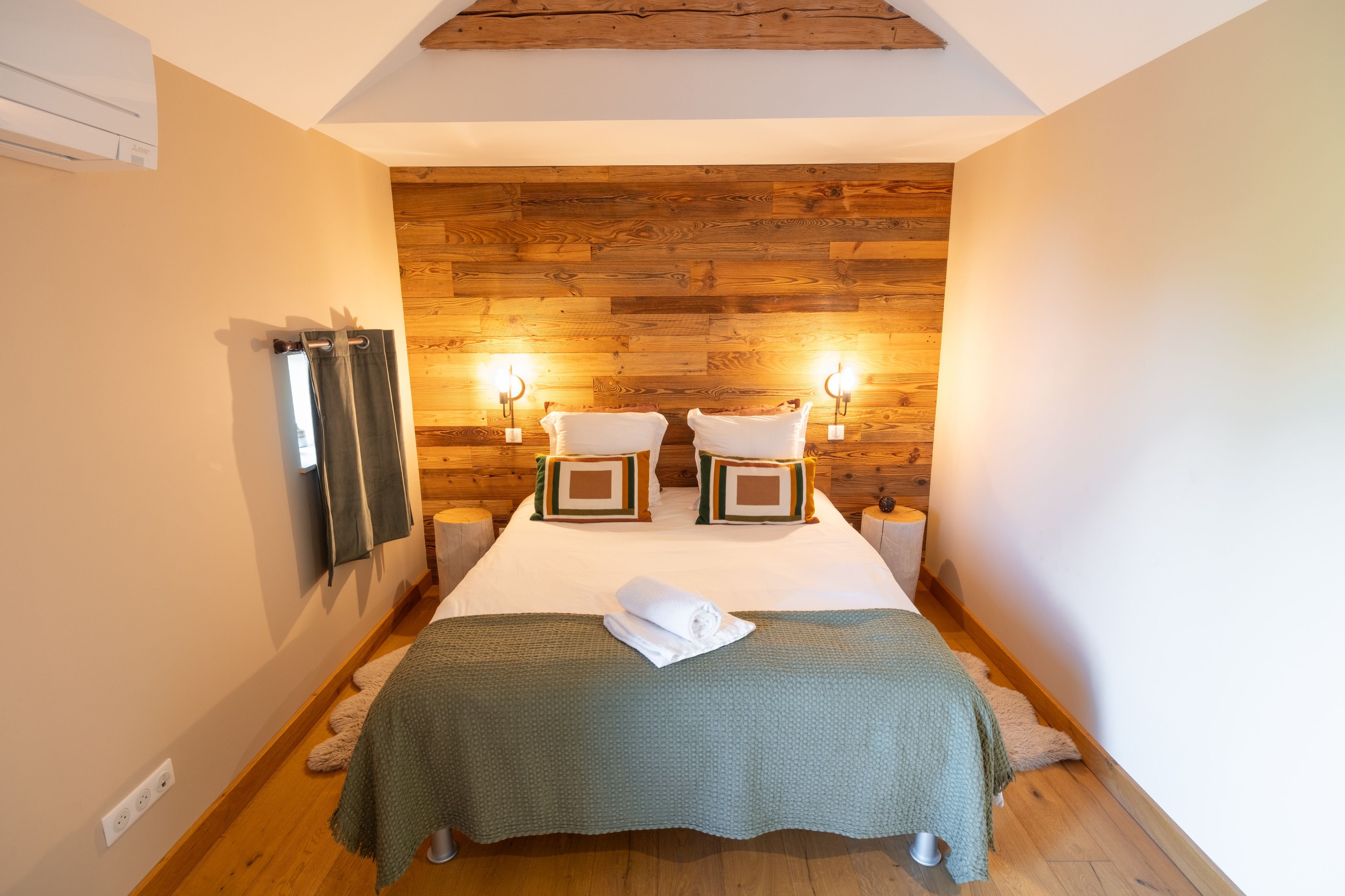 Ett smalt hotellrum med en väggpanel av trä, två sänglampor, en säng med ett grönt överkast och ett litet nattduksbord.