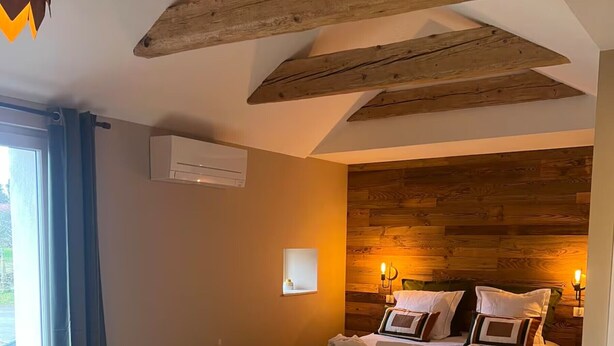 Une chambre avec un plafond en bois, un lit avec des oreillers et une couverture, un tapis blanc et moelleux, et une fenêtre avec des rideaux.