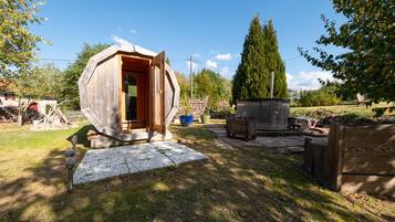 A wooden cabin with an open door, surrounded by a garden with a stone pathway, a tall tree, and a metal cart.