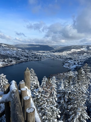 A panoramic view of a snow-covered landscape with a lake, forested hills, and a wooden fence in the foreground.