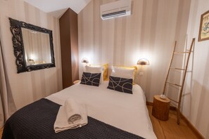 A neatly made bed with a dark blanket and pillows, a wall-mounted mirror, a bedside lamp, and a small wooden stool. - Guest Room “Épicéa” with Private Bathroom, Shared Terrace, Wi-Fi & Air Conditioning (Champdray)