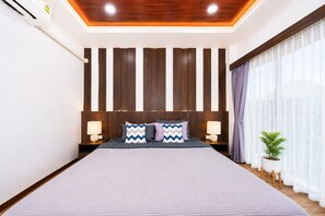 A bedroom with a large bed, wooden panel wall, two bedside tables with lamps, a potted plant, and a view of a building outside.