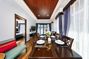 Habitación de hotel con comedor, sofá, televisor y un amplio ventanal con cortinas.