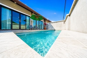 A swimming pool with clear water, a lounge area with chairs, and a large windowed room with curtains.