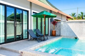 Una piscina con lettini da relax e un ombrellone, una pianta in vaso e un edificio con ampie vetrate e facciata rivestita di piastrelle.