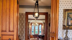 Um corredor de madeira com um candelabro, uma área de jantar com cadeiras e uma mesa, e uma lareira com uma estátua.