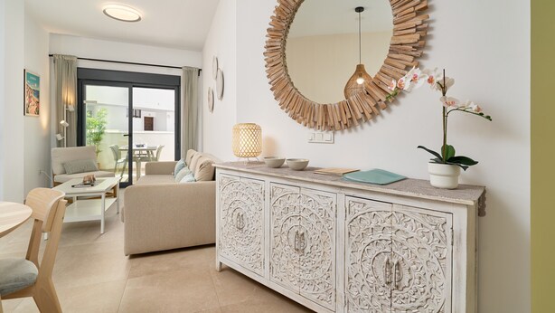 A modern living room with a white sofa, a dining table, a white cabinet with intricate carvings, a round mirror with a wooden frame, and a potted orchid plant. - Apartment 'Building Copenhagen, Nerja' with Shared Pool, Wi-Fi and Air Conditioning (Nerja)