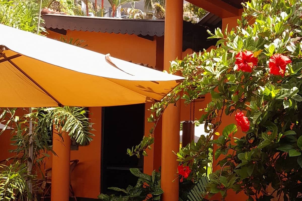 An outdoor seating area with wicker chairs and a yellow umbrella, surrounded by greenery and a building with orange walls and a tiled roof.