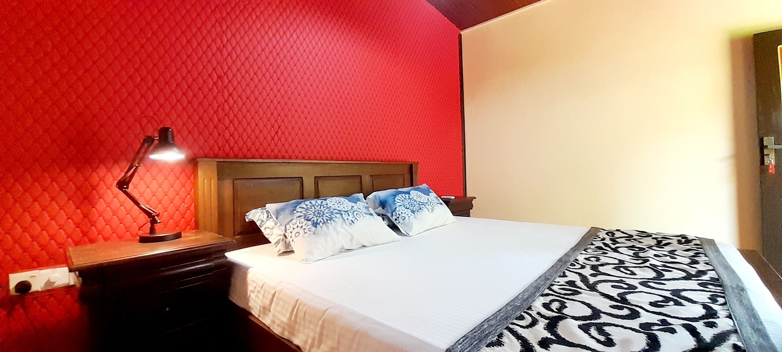 A bedroom with a red textured wall, a wooden headboard, a white bed with patterned bedding, and a nightstand with a lamp.