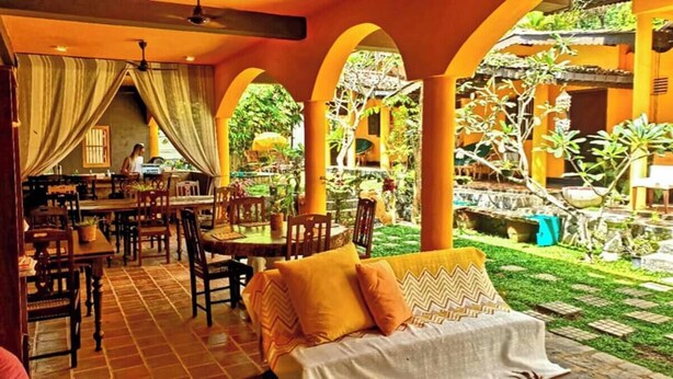 An indoor dining area with tables and chairs, a sofa with cushions, and a view of a garden with a water feature. - The Fat Mermaid (Mirissa)