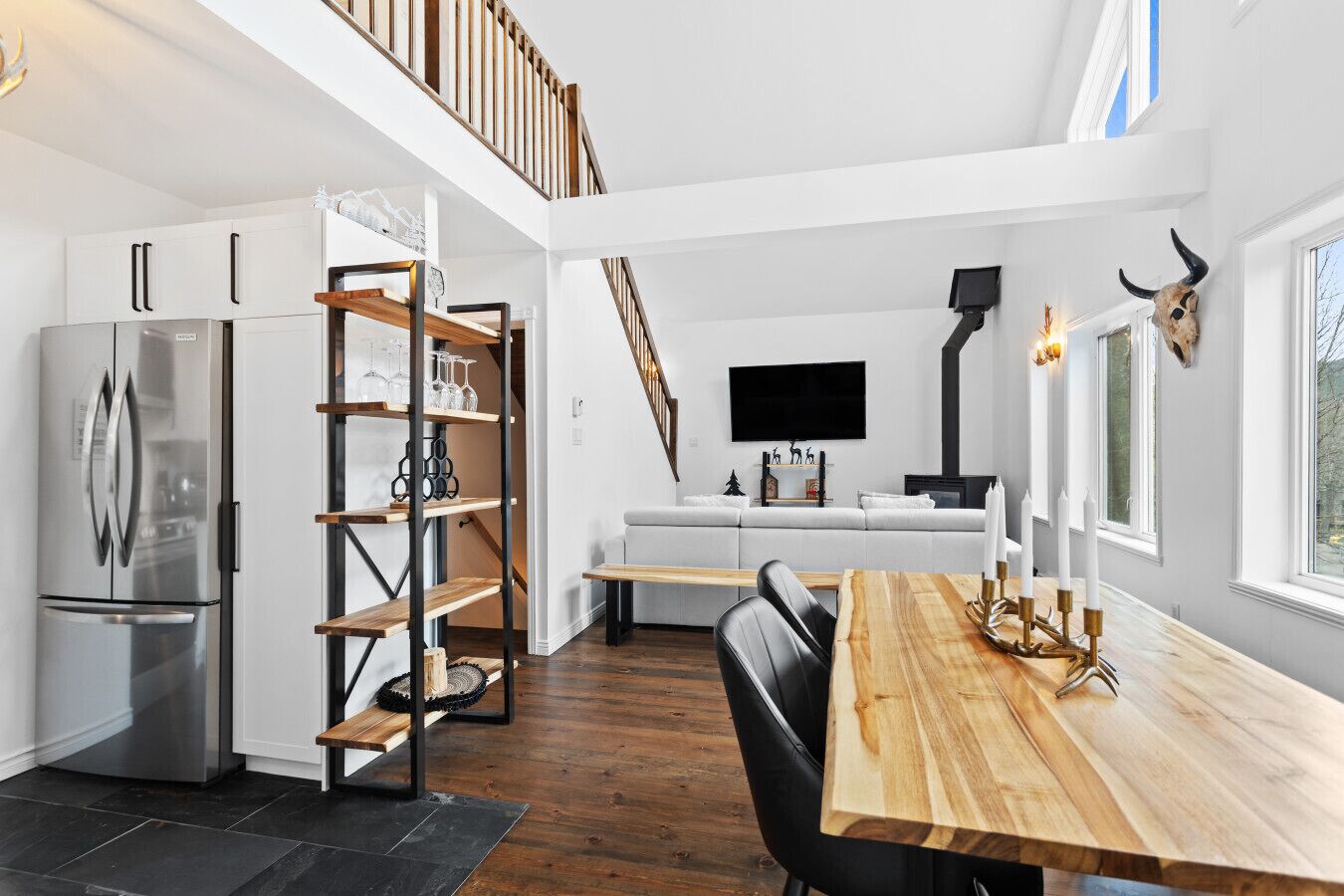 Une cuisine moderne dotée d’un réfrigérateur encastré, d’étagères en bois, d’une table à manger décorée de bougies et d’un escalier menant à un niveau supérieur.