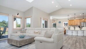 A spacious living room with a white sofa, a coffee table, and a kitchen area with bar stools. - Serene coastal ambiance, fenced yard, fire pit, and outdoor furniture! (Seaside)