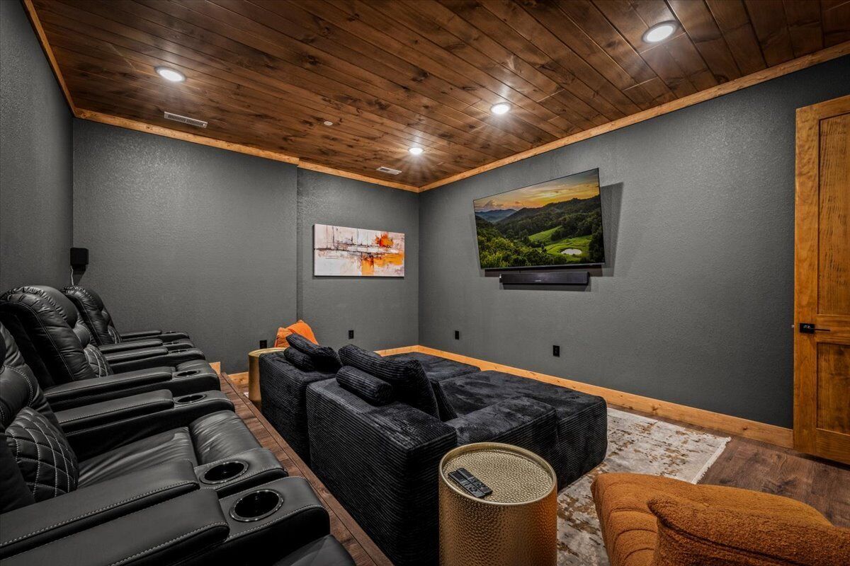 Sala de cine en casa con pared gris oscuro, techo de madera, asientos reclinables de cuero negro, un sofá negro, un televisor de pantalla plana grande y un cuadro de un paisaje.