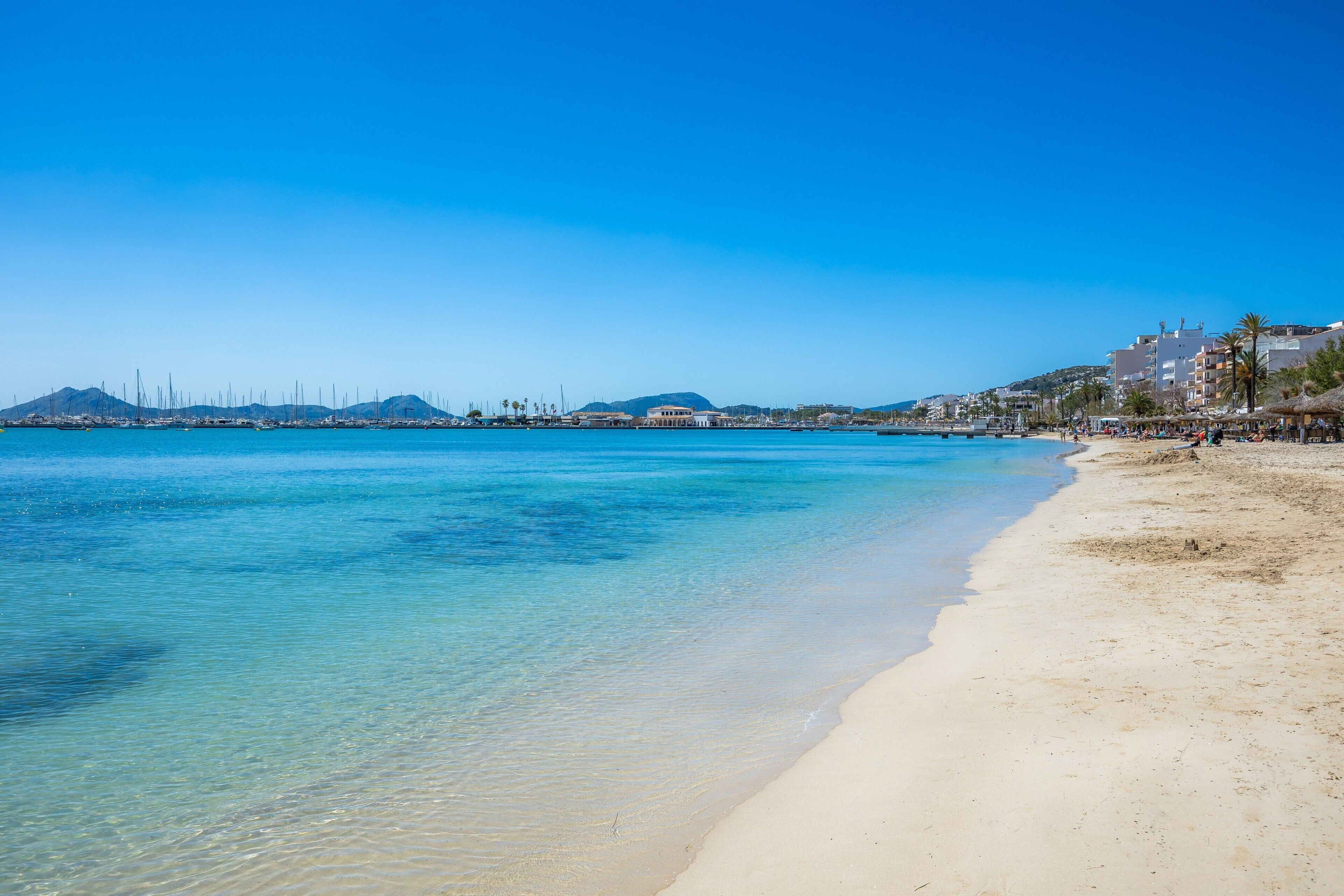 A beach with clear water, a sandy shore, and buildings in the background.