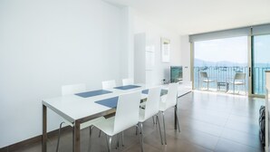 Une salle à manger moderne avec une grande table, des chaises blanches et une vue sur la mer, visible à travers une porte vitrée.