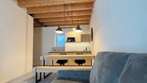 A modern living room with a wooden ceiling, a grey sofa, and a kitchen area in the background.